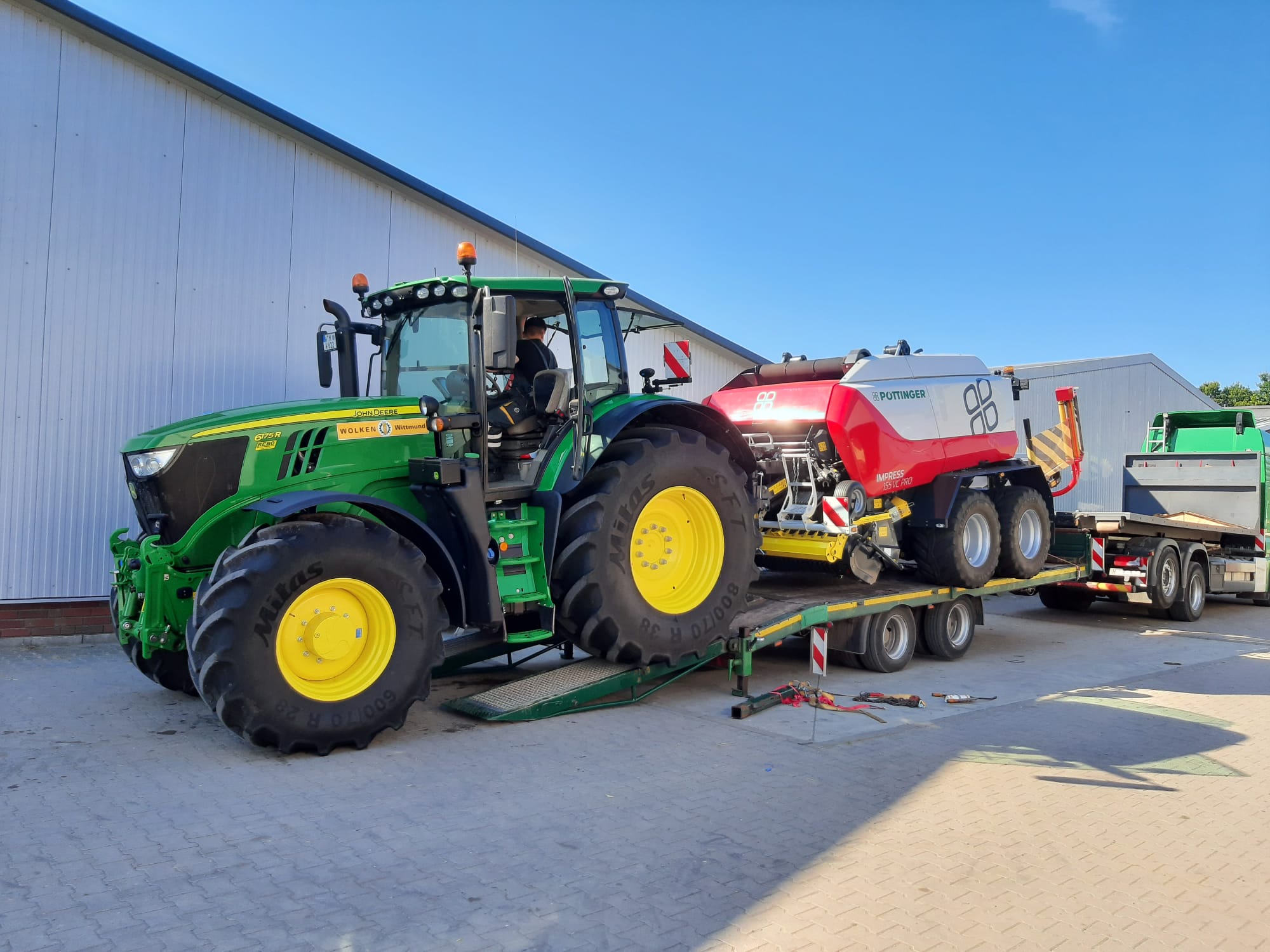 John Deere Traktor lädt Rundballenpresse auf Tieflader vor Hofgebäuden.