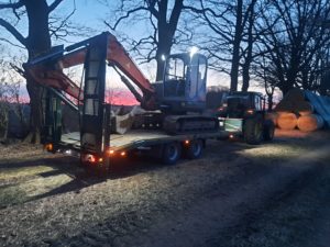 Kompaktbagger auf Tieflader, Traktor zieht über Waldweg, Sonnenuntergang und Rundballen.