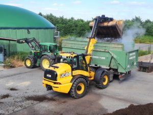 Gelber JCB-Teleskoplader belädt Brantner Anhänger mit dampfendem Kompost, Biogasanlage.
