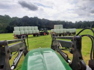 Zwei Traktoren ziehen Anhänger mit grünen Silageballen über frisch gemähte Wiese.