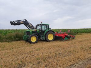 Grüner Traktor mit Frontlader und Scheibenegge auf Stoppelfeld vor Maisfeld, bewölkt.