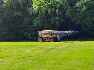 Gelber Gülle-Selbstfahrer mit Schleppschuhverteiler auf grüner Wiese am Waldrand, sonnig.