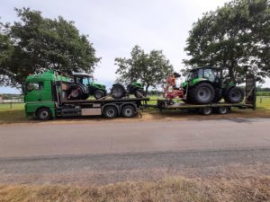 MAN-Lkw mit drei Deutz-Traktoren auf ländlicher Straße, Bäume und Weiden im Hintergrund.
