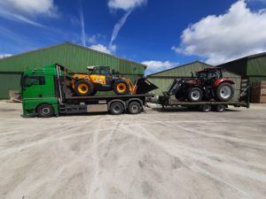 MAN-Tieflader mit gelbem JCB-Teleskoplader und rotem Case-Traktor vor grünen Hallen.