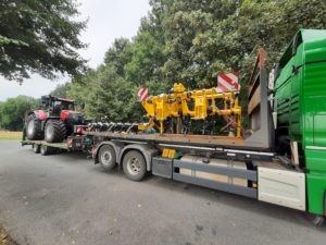 Grüner MAN-Tieflader transportiert gelbes Bodenbearbeitungsgerät und roten Traktor am Waldrand.