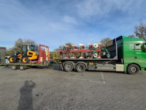 Grüner Lkw transportiert gelben JCB Frontlader und rot-grünen Pflug, Parkplatz, klarer Himmel