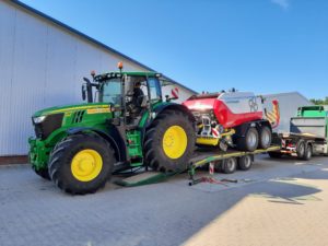Grüner John Deere Traktor lädt Pöttinger Rundballenpresse auf Tieflader, Gebäude im Hintergrund.