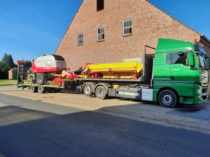 MAN-Sattelzug transportiert Pöttinger Rundballenpresse und Mähwerk vor Ziegelgebäude.