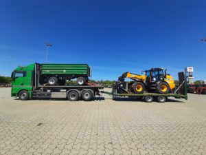 Grüner Lkw zieht Tieflader mit grünem Anhänger und gelbem JCB-Teleskoplader auf Platz.