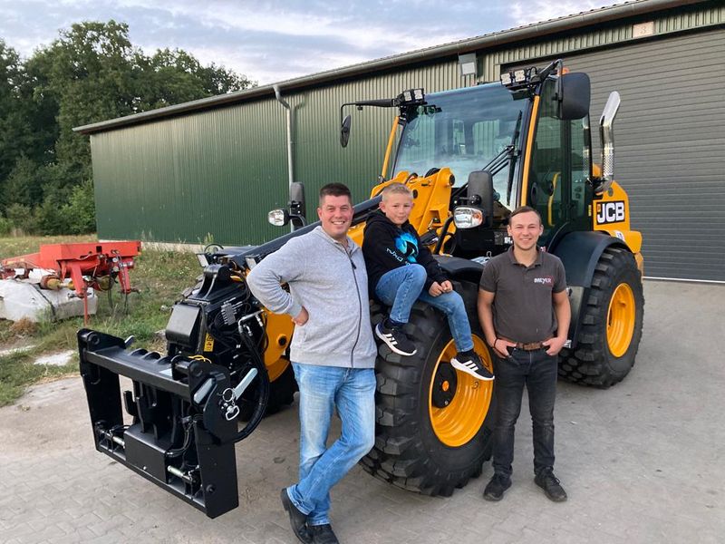 Drei Personen vor neuem JCB-Teleskoplader bei Maschinenhalle mit grünem Dach.